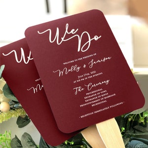 May include: Burgundy wedding ceremony program fans with white calligraphy that reads "We Do" and "Molly & Jamison". The program lists the ceremony details, including the date, time, and order of events.