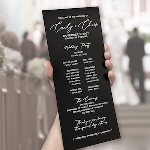 May include: A black wedding program with white calligraphy text. The program lists the wedding party, ceremony details, and a thank you message. The program is held in a person's hand, with a blurred background.