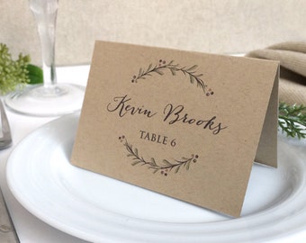 Op de afbeelding: Een bruine plaatskaart met de handgeschreven naam "Kevin Brooks" en de tekst "Tafel 6". De kaart is versierd met een krans van bladeren en bessen.