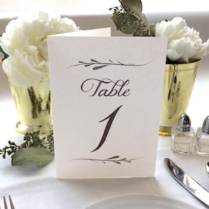 May include: White card with brown calligraphy that reads "Table 1" with a simple floral design. The card is on a white tablecloth with white flowers in gold vases and salt and pepper shakers.