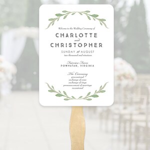 May include: A white wedding fan with a wooden handle. The fan features text announcing the wedding of Charlotte and Christopher, with the date and location. Green watercolor leaf accents frame the text.