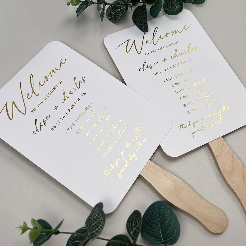 May include: Two white wedding fans with gold lettering. The fans feature the text "Welcome to the wedding of Elise + Charles 08.17.24 | Austin, TX" and a wedding timeline.