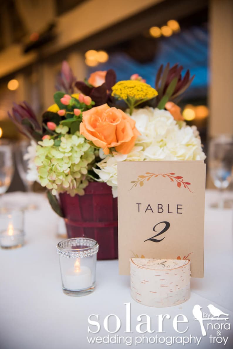 May include: A table number card with the text "TABLE 2" on a birch wood base. The card is decorated with a floral design in shades of orange and red. The card is sitting on a white table with a vase of flowers in the background.