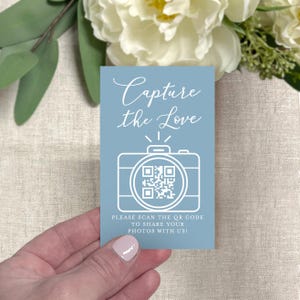May include: A light blue card with the text "Capture the Love" in cursive script. Below the text is a white camera graphic with a QR code. The card also includes the text "PLEASE SCAN THE QR CODE TO SHARE YOUR PHOTOS WITH US!"