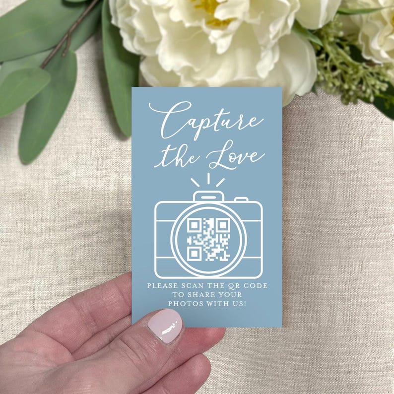 May include: A light blue card with the text "Capture the Love" in cursive script. Below the text is a white camera graphic with a QR code. The card also includes the text "PLEASE SCAN THE QR CODE TO SHARE YOUR PHOTOS WITH US!"