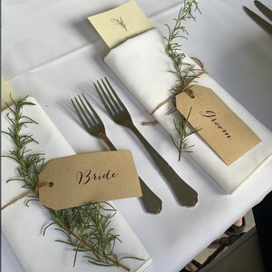 May include: Two place settings for a wedding reception with white napkins, rosemary sprigs, and brown paper place cards that say "Bride" and "Groom".