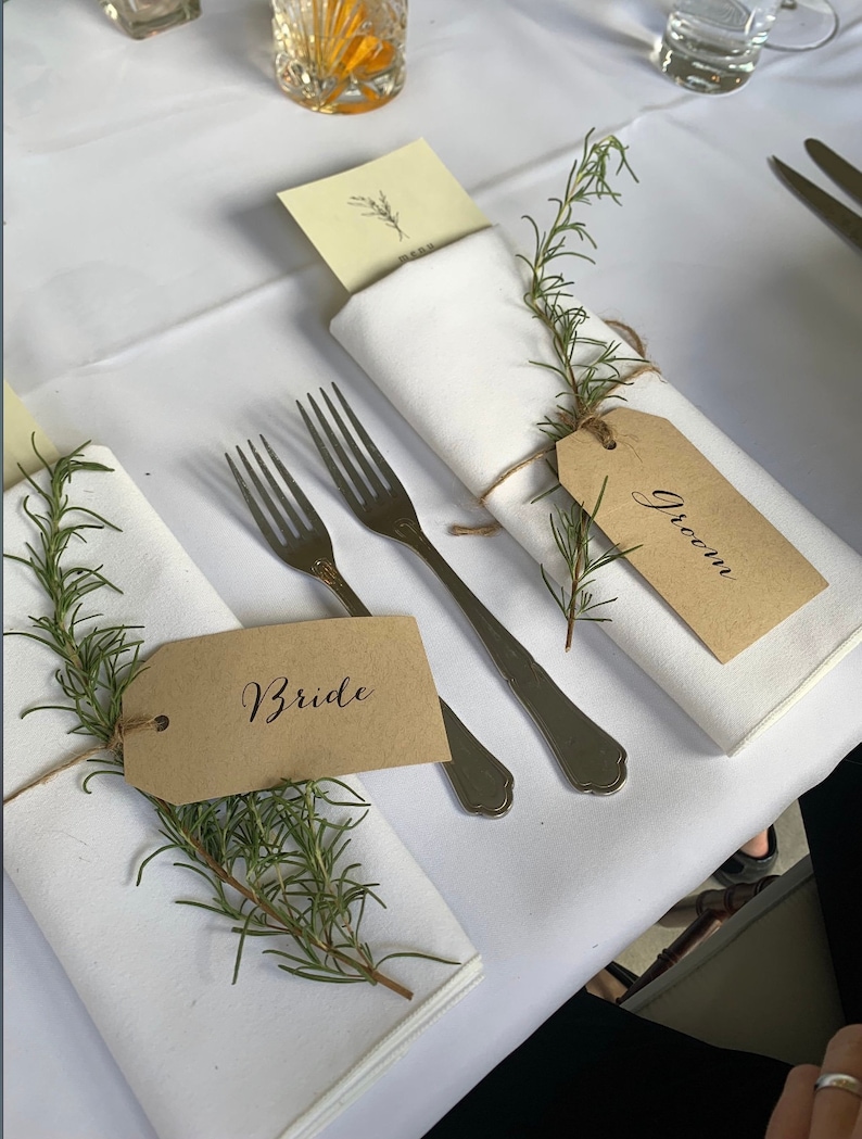 May include: Two place settings for a wedding reception with white napkins, rosemary sprigs, and brown paper place cards that say "Bride" and "Groom".