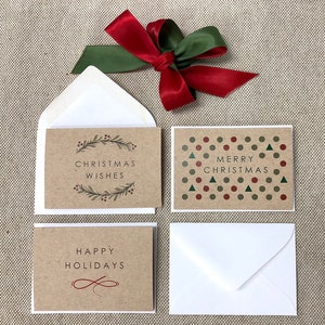 May include: Set of three Christmas cards with brown paper cardstock and white envelopes. The cards feature different holiday greetings: "Christmas Wishes" with a wreath design, "Merry Christmas" with a pattern of red circles and green triangles, and "Happy Holidays" with a simple infinity symbol.