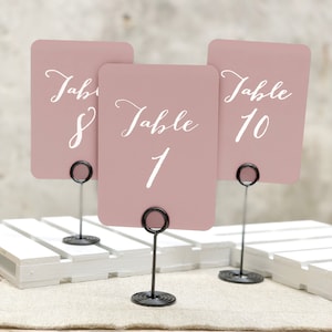 May include: Three blush-colored table number cards with white calligraphy text, reading "Table 1", "Table 8", and "Table 10". Each card is held by a black metal wire stand with a circular base. The cards are displayed on a white wooden pallet.