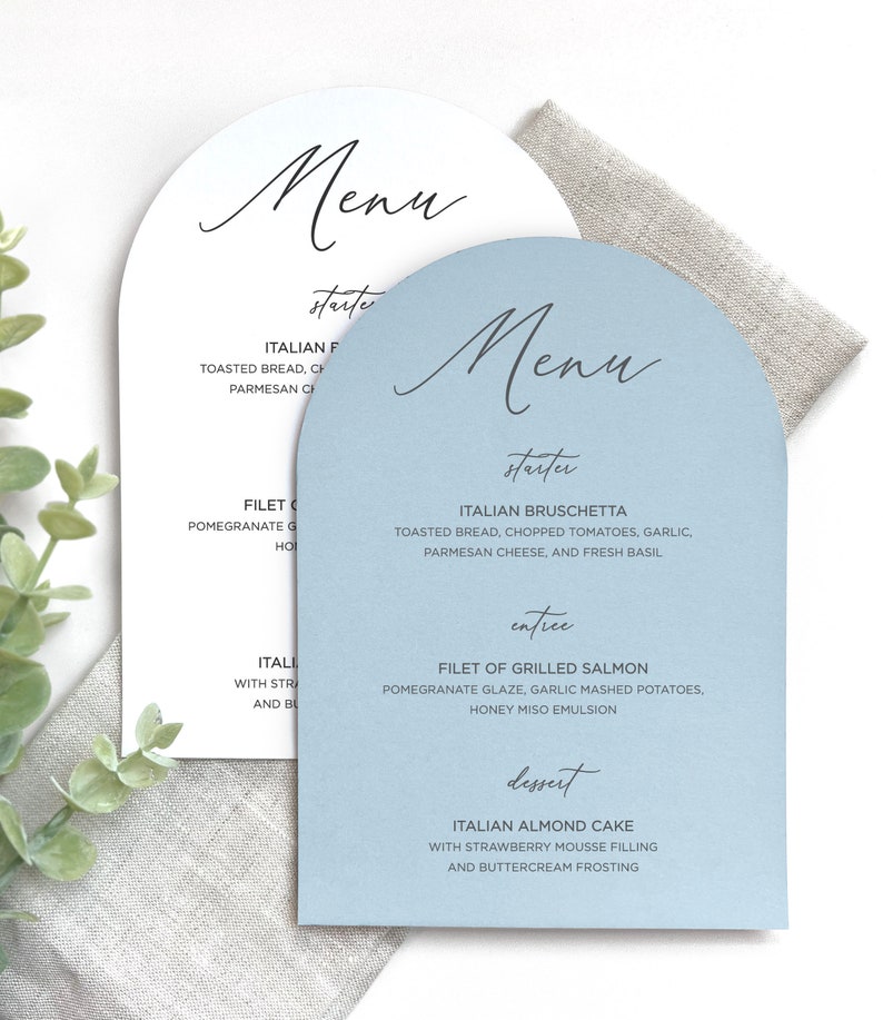 May include: Two arch-shaped menu cards, one white and one light blue, featuring black text. The blue menu lists Italian Bruschetta, Filet of Grilled Salmon, and Italian Almond Cake. The text is printed in a combination of script and sans-serif fonts.