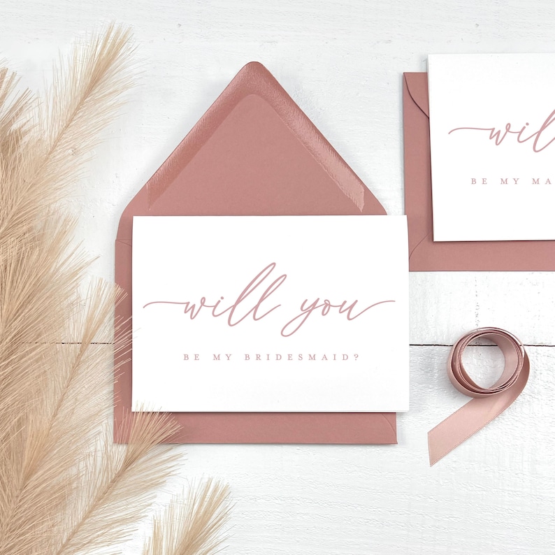 May include: A white card with the text "will you BE MY BRIDESMAID?" in a soft pink script is placed inside a matching pink envelope. A second card and envelope are visible, along with a rolled pink ribbon and dried decorative grass.