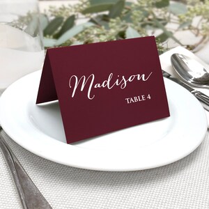 May include: A burgundy place card with the name "Madison" in white script and "TABLE 4" printed below. The card is on a white plate, with silverware and a napkin visible in the background. The setting suggests a formal event.