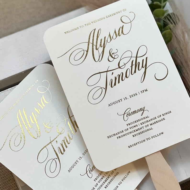 May include: Two off-white wedding ceremony program fans with rounded corners and gold foil lettering. The text reads: Welcome to the wedding ceremony of Alyssa & Timothy, August 15, 2026, 5PM, Ceremony, Processional, Exchange of Vows, Exchange of Rings, Pronouncement of Marriage, Recessional, Reception to follow.