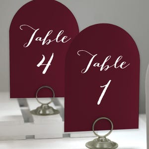 May include: Two burgundy table number cards with arched tops. The cards have white cursive text that reads "Table" and a number. Table 4 and Table 1 are visible. The cards are held by silver metal holders.