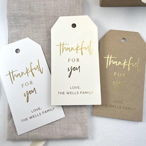 May include: Three gift tags with the text "thankful for you" in gold foil lettering. The tags are on a beige linen background. The bottom of each tag has the text "Love, The Wells Family".