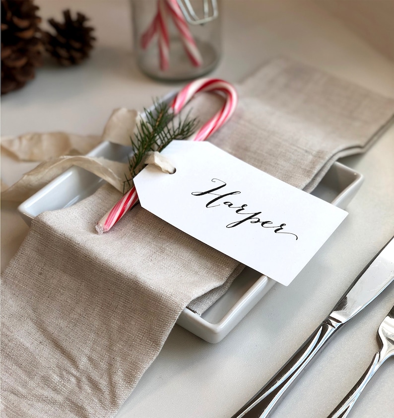 May include: A white gift tag with the name "Harper" written in black calligraphy, tied to a red and white candy cane with a sprig of green pine. The tag is placed on a folded linen napkin, next to a white square plate and silver cutlery.