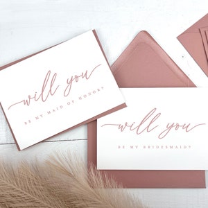 May include: White cards with the text "will you" in a delicate script, with the phrases "BE MY MAID OF HONOR?" and "BE MY BRIDESMAID?" in a matching color. The cards are set on a white surface with mauve envelopes.