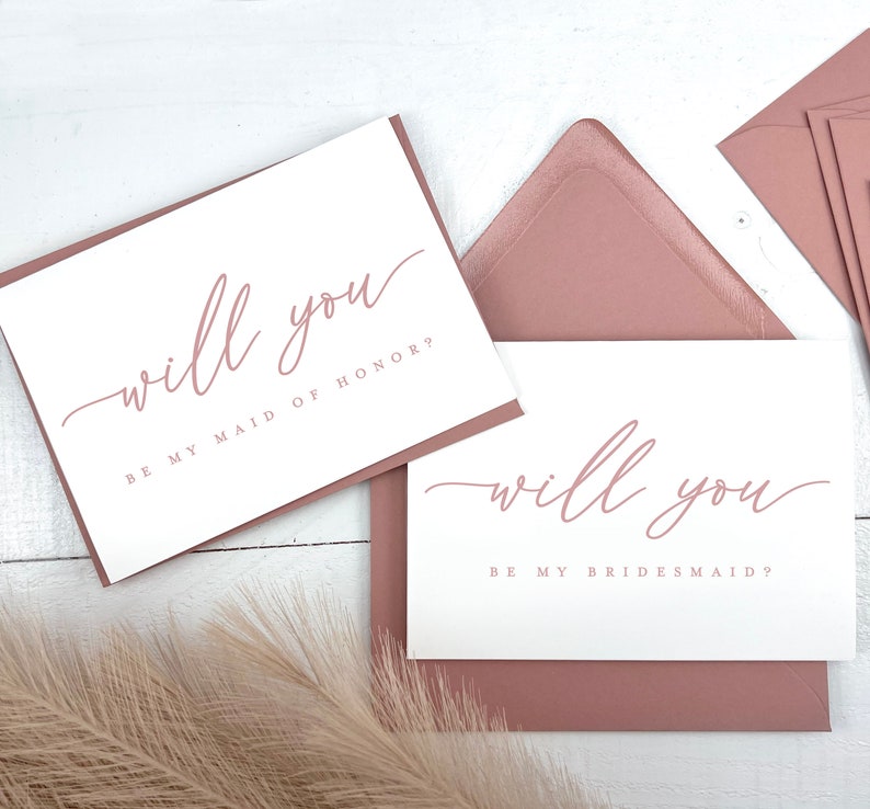 May include: White cards with the text "will you" in a delicate script, with the phrases "BE MY MAID OF HONOR?" and "BE MY BRIDESMAID?" in a matching color. The cards are set on a white surface with mauve envelopes.