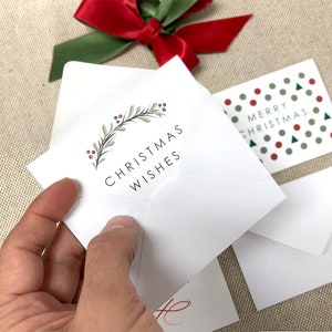 May include: A white envelope with a green wreath and red berries design. The text "Christmas Wishes" is printed in black on the envelope.