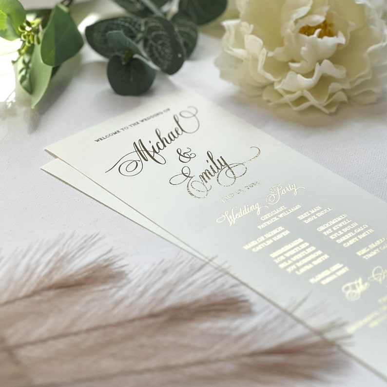 May include: A cream-colored wedding program card featuring gold foil lettering. The text reads Welcome to the wedding of Michael & Emily, July 28, 2024, and lists the wedding party members including the officiant, maid of honor, and groomsmen.