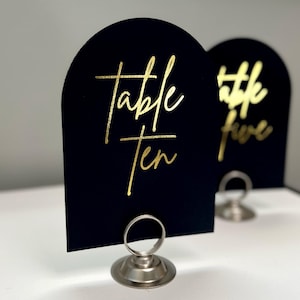May include: Black table number card with gold lettering that reads "Table Ten" on a silver stand.