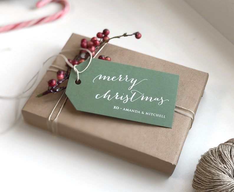 May include: A rectangular gift box wrapped in brown paper with a green gift tag attached by white string. The tag features white text that reads merry christmas and XO - AMANDA & MITCHELL. A small sprig of red berries is tucked under the string.