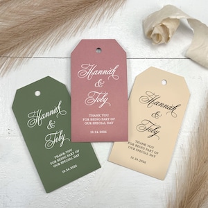 May include: Three wedding favor tags with the names Hannah & Toby, written in cursive script. The tags are in different colors: green, pink, and cream. Each tag has the text "Thank you for being part of our special day" and the date "10.24.2026".
