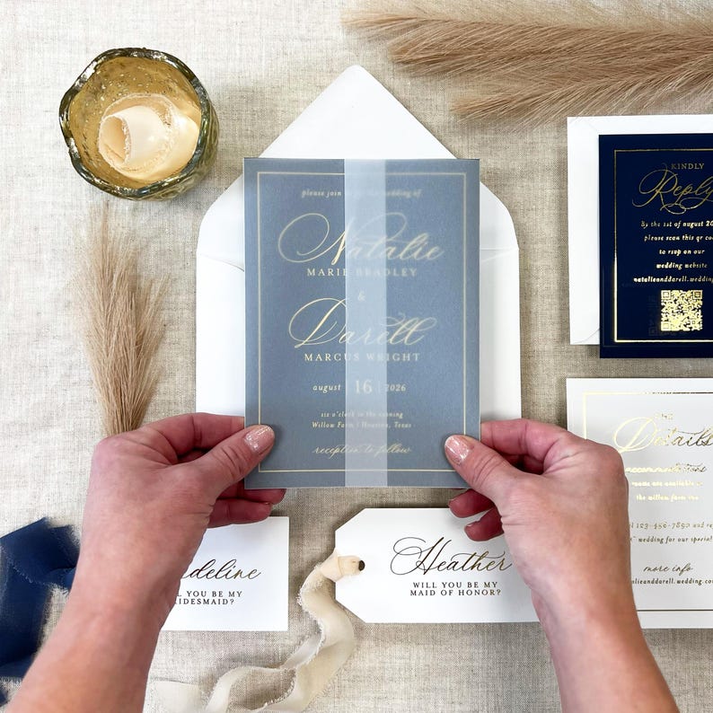 May include: A wedding invitation suite featuring a blue and gold color scheme. The main invitation card is held by hands and includes the names "Natalie" and "Darell". Other cards include RSVP and details cards. Decorative elements include pampas grass and a gold candle holder.