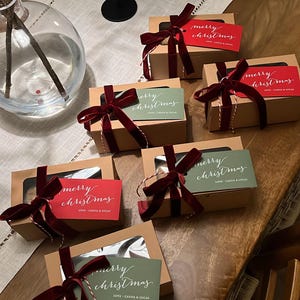 May include: Five rectangular cardboard gift boxes with clear windows, each tied with a dark red velvet ribbon and a red or green tag reading "merry christmas" and "LOVE - CASSIA & LUCAS" in white script, arranged on a wooden table.