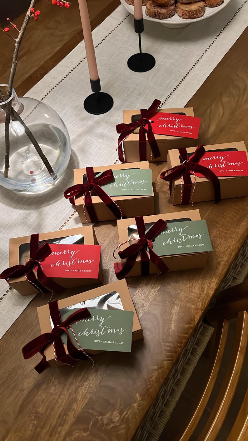 May include: Five rectangular cardboard gift boxes with clear windows, each tied with a dark red velvet ribbon and a red or green tag reading "merry christmas" and "LOVE - CASSIA & LUCAS" in white script, arranged on a wooden table.