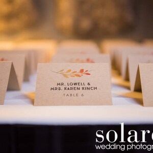 May include: A set of table cards with fall-themed foliage designs. The cards are printed with the names of guests and their assigned table numbers. The text on the cards reads "Dennis & Megan Ebert, Table 7", "Mr. Lowell & Mrs. Karen Kinch, Table 6", and "Mr. Paul & Mrs. Sheryl, Table ".