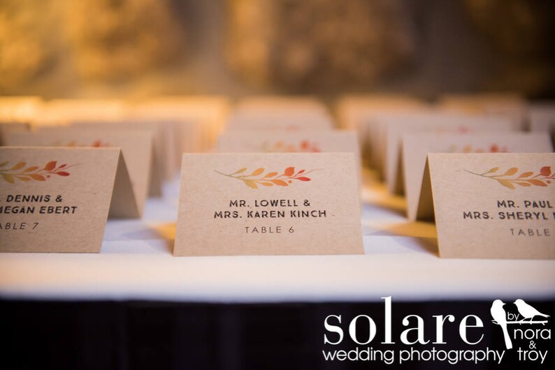May include: A set of table cards with fall-themed foliage designs. The cards are printed with the names of guests and their assigned table numbers. The text on the cards reads "Dennis & Megan Ebert, Table 7", "Mr. Lowell & Mrs. Karen Kinch, Table 6", and "Mr. Paul & Mrs. Sheryl, Table ".