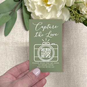 May include: A sage green card with the text "Capture the Love" in elegant script. A white outline of a camera with a QR code is below the text. The card also includes the text "PLEASE SCAN THE QR CODE TO SHARE YOUR PHOTOS WITH US!"