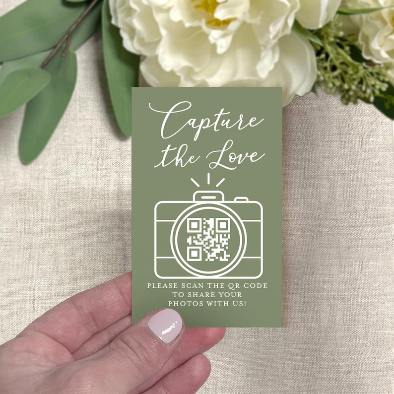 May include: A sage green card with the text "Capture the Love" in elegant script. A white outline of a camera with a QR code is below the text. The card also includes the text "PLEASE SCAN THE QR CODE TO SHARE YOUR PHOTOS WITH US!"