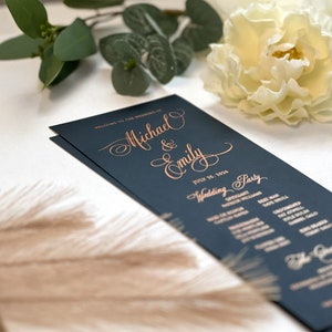 May include: A navy blue wedding program with gold lettering. The program lists the names "Michael & Emily" and the date "July 25, 2016". The program also includes a list of wedding party members.