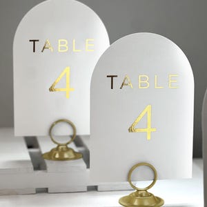 May include: Two white table number cards with gold foil lettering. The cards are shaped like arches and have a gold metal stand. The cards read "Table 4".