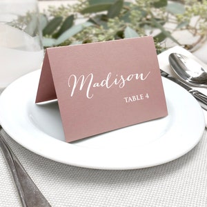 May include: A folded, dusty rose place card with the name "Madison" in elegant white script and "TABLE 4" printed below. The card rests on a white plate, with silverware and a napkin in the background, suggesting a formal dining setting.