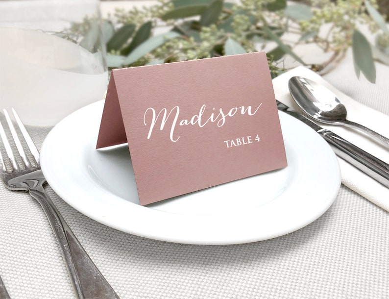 May include: A folded, dusty rose place card with the name "Madison" in elegant white script and "TABLE 4" printed below. The card rests on a white plate, with silverware and a napkin in the background, suggesting a formal dining setting.