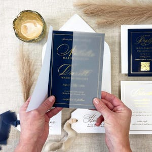 May include: Navy blue wedding invitation with gold script, held open to reveal the details. The invitation includes a translucent overlay. Additional stationery includes a reply card, details card, and a tag with a ribbon.