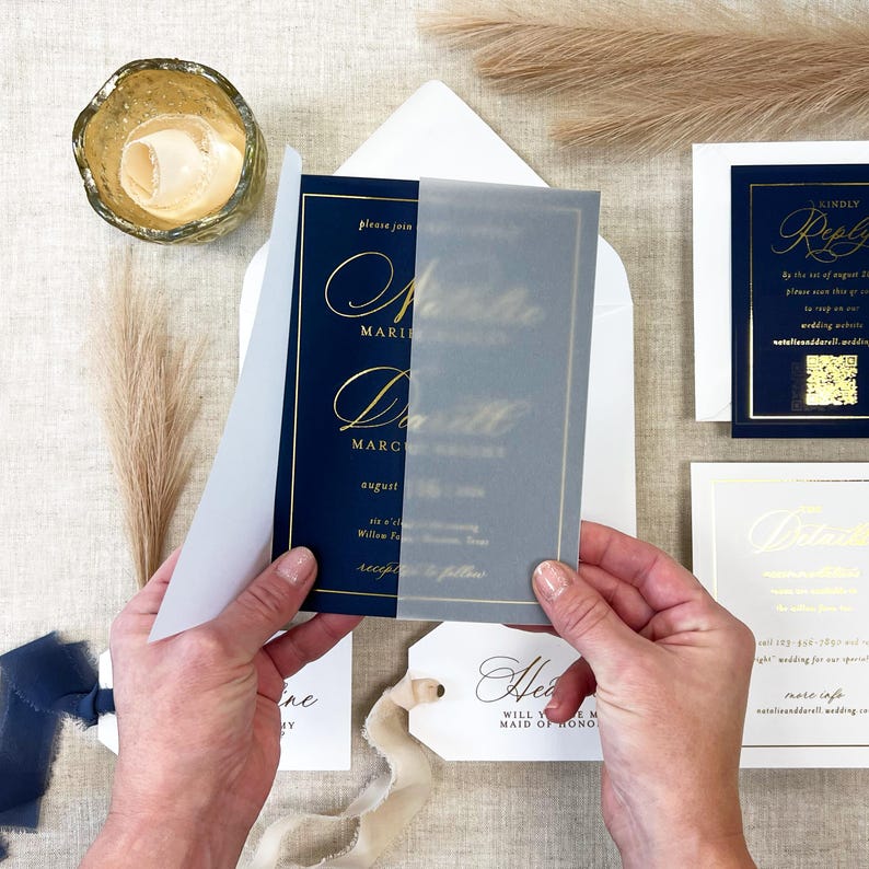 May include: Navy blue wedding invitation with gold script, held open to reveal the details. The invitation includes a translucent overlay. Additional stationery includes a reply card, details card, and a tag with a ribbon.
