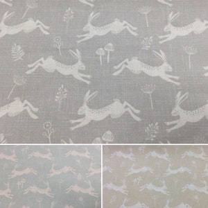 Op de afbeelding: Een stof met een herhalend patroon van witte konijnen die over een grijze achtergrond rennen. De stof is verkrijgbaar in drie kleuren: grijs, lichtblauw en beige.