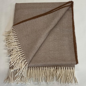 May include: A folded throw blanket featuring a light brown and white herringbone pattern. The blanket has a solid dark brown border along the edges and is finished with long, cream-colored fringe tassels along the bottom and side edges.