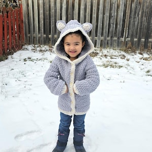 May include: A young child wearing a light purple crocheted hooded cardigan with white trim and ear accents. The cardigan has a button closure. The child is standing in the snow.