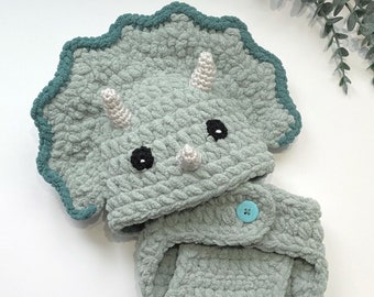 May include: A crocheted baby outfit in the shape of a triceratops dinosaur. The outfit is green with a blue trim and has a button closure.