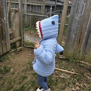 May include: A light blue crocheted shark hoodie with a white and red mouth and a black eye. The hoodie is being worn by a child standing in a garden.