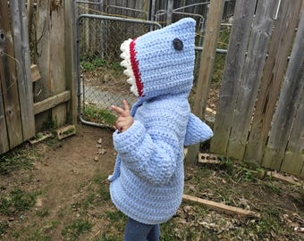 Puede incluir: Una sudadera con capucha de tiburón de crochet azul claro con una boca blanca y roja y un ojo negro. La sudadera la lleva puesta un niño de pie en un patio.