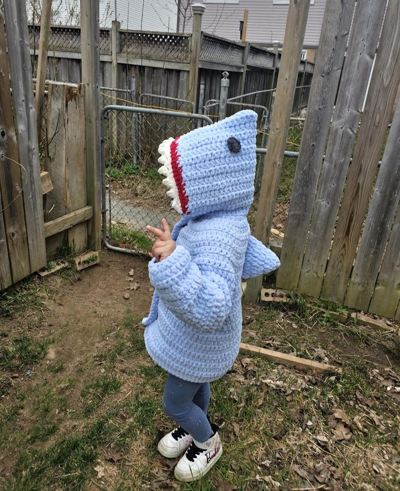 Puede incluir: Una sudadera con capucha de tibur&oacute;n de crochet azul claro con una boca blanca y roja y un ojo negro. La sudadera la lleva puesta un ni&ntilde;o de pie en un patio.