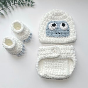 May include: A white crocheted baby outfit with a snowman design. The outfit includes a hat, booties, and a diaper cover. The snowman has a blue face with black eyes and a black mouth.