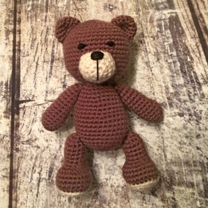 Puede incluir: Un oso de peluche de crochet marr&oacute;n con la cara blanca y los ojos negros. El oso est&aacute; de pie sobre una superficie de madera.