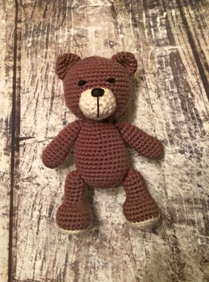 Puede incluir: Un oso de peluche de crochet marr&oacute;n con la cara blanca y los ojos negros. El oso est&aacute; de pie sobre una superficie de madera.